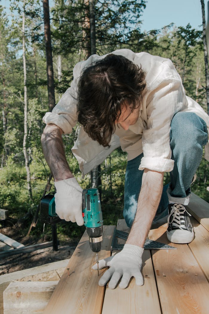 A worker operates a drill on a wooden deck in the forest, captured in daylight.